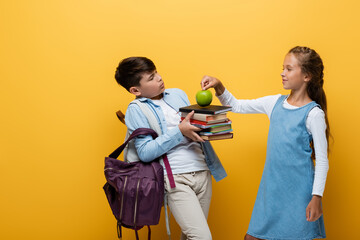 Wall Mural - Schoolgirl putting apple on books near asian friend on yellow background.