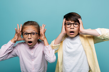 Wall Mural - Scared interracial schoolchildren in eyeglasses looking at camera isolated on blue.