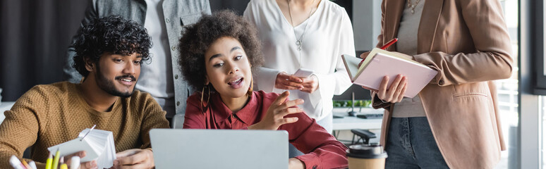 Wall Mural - african american woman talking near laptop and indian colleague in advertising agency, banner.