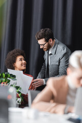 Wall Mural - happy african american woman holding papers near man in eyeglasses and blurred colleague in ad agency.