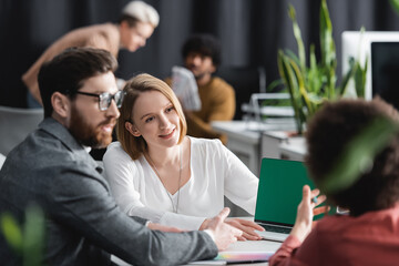 Wall Mural - woman smiling near multiethnic colleagues and laptop with green screen in ad agency.