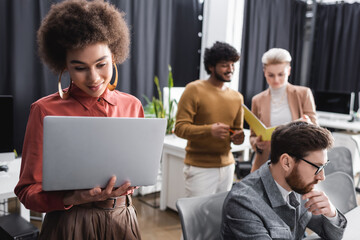 Wall Mural - positive african american woman looking at laptop near multiethnic colleagues working in ad agency.