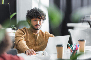 young indian designer working near laptop in advertising agency on blurred foreground.