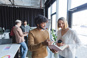 Wall Mural - smiling indian man pointing at notebook near colleague in advertising agency.