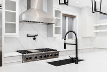 A Luxury Kitchen Detail Shot With A Black Faucet, White Cabinets And Marble Countertops, And Stainless Steel Appliances.