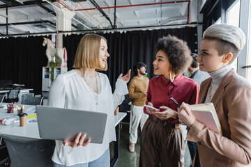 Wall Mural - smiling multicultural women with gadgets and notebook talking in advertising agency.