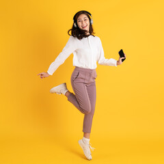 Wall Mural - Cute young asian teenage woman using smartphone wearing headphone isolated on yellow background jumping and dancing