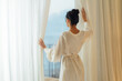 © Alla - Morning of beautiful young woman opening curtains in bedroom and enjoying of the amazing views. Back view of the young lady