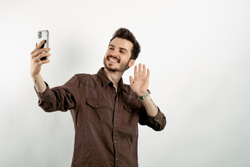 Wall Mural - Cheerful young man wearing brown shirt posing isolated over white background taking selfie or having video call and waving hand. Recording vlog, social media influencer streaming, making video call.