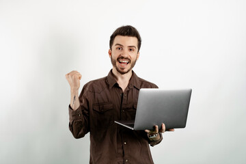 Wall Mural - Cheerful caucasian man wearing brown shirt posing isolated over white background using laptop computer while making winner gesture.