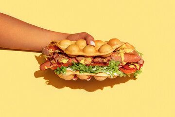 Wall Mural - Hand holding bubble waffle hot dog with sausage, tomato, lettuce, bacon, sauce, mustard, cheese, red bell pepper isolated on yellow background. Fast food, take away. Snack