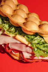 Wall Mural - bubble waffle sandwich with ham, tomato, lettuce, bacon, sauce, mustard, cheese, red bell pepper isolated on red background. Fast food, take away. Snack. Macro