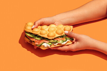 Canvas Print - Hand holding bubble waffle sandwich with salmon, arugula, lettuce, cream cheese and cucumber  isolated on orange background. Fast food, take away. Snack