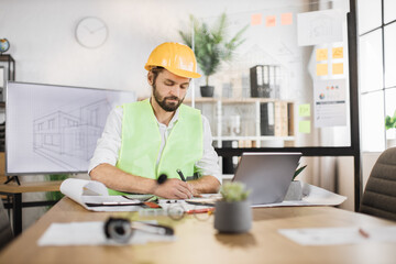 Young builder, engineer in reflective vest uniform and hard hat on desk in office on background of large TV screen with , browsing in laptop and looking through papers or sketches.