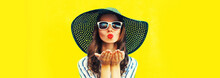 Portrait Of Beautiful Young Woman Blowing Her Lips Sending Air Kiss Wearing Sunglasses, Summer Straw Hat On Yellow Background