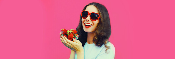 Portrait close up of happy smiling woman with handful of fresh strawberries wearing red heart shaped sunglasses on pink background