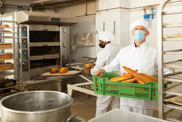 Wall Mural - Baker in face mask for viral protection holding crate with bread, industrial kitchen of bakery on background