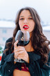 Portrait of young sexy woman with wavy dark hair, make-up wearing unbuttoned black shirt, red bra, standing on balcony, holding glass with red wine, licking red lips. Celebration, New year, holiday. 