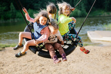 Four Children Swinging On Wicker Swing On Playground In Park By Lake, Sand And Children's Summer Vacation With Friends