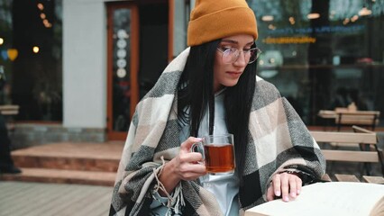 Wall Mural - Smiling young brunette woman wearing hat and eyeglasses reading book and drinking tea in cafe outdoors