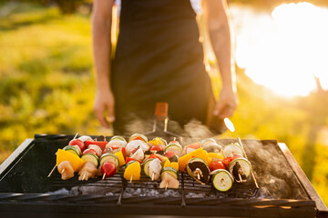 Wall Mural - Crop man roasting vegetable kebabs on grill near lake