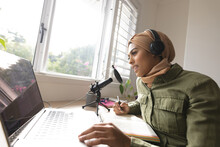 Smiling Biracial Young Woman In Hijab Recording Podcast Through Microphone At Home Studio