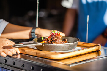 Wall Mural - Waiter hands holding wooden tray with fresh cooked meat in a frying pan on wooden table in luxury restaurant. Grilled beaf with vegetables and chef on background. Fine dining food, food serving.