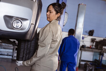 Mid Adult Asian Female Mechanic Fixing Car's Tire In Workshop With Colleague In Background