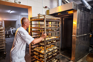 Professional baker in uniform inserts cart with decks for baking raw dough to make bread in an industrial oven in a bakery