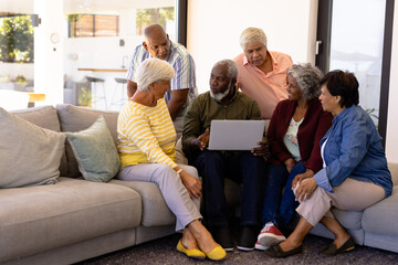 Wall Mural - Multiracial senior man holding laptop explaining to friends while sitting on sofa in nursing home