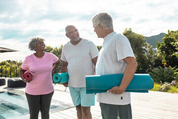 Wall Mural - Happy multiracial senior friends with exercise mats talking while standing against sky in yard