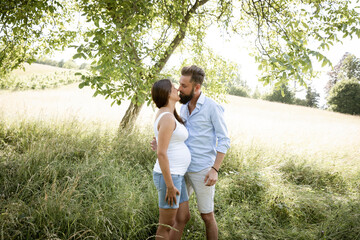 Wall Mural - pretty young pregnant woman with white shirt stands with her boyfriend with beard and blue shirt in high flower meadow and cuddle