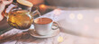 Banner of grey cup with teapot. The hand holding the teapot pours herbal tea into pot on saucer in soft focus on blurred background. Coffee, tea house, bokeh lights. The concept of cozy pastime
