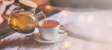 Banner Of Grey Cup With Teapot. The Hand Holding The Teapot Pours Herbal Tea Into Pot On Saucer In Soft Focus On Blurred Background. Coffee, Tea House, Bokeh Lights. The Concept Of Cozy Pastime