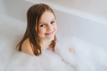 a small, smiling, beautiful red-haired girl with long hair, the child bathes, washes in a white bath