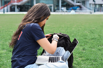 Wall Mural - Young girl student sitting on green grass lawn on campus with laptop computer studying