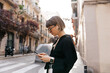 Outside profile portrait of stylish pretty girl with short hairstyle wearing black shirt in wireless headphones scrolling smartphone and enjoying walking on summer street