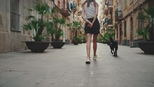 Girl Walks Along Narrow Street In The Old Town With Her Black Dog. Back View
