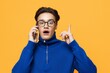 © Tatiana - a cute, joyful man stands on an orange background in a blue zip-up jacket and black eyeglasses, talking on the phone and pointing at him with his finger