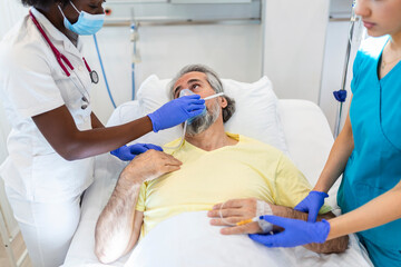 Wall Mural - Young doctor and nurse wearing a surgical mask checking on a senior male patient wearing a positive pressure oxygen mask to aid breathing in a hospital bed during the Covid-19 pandemic