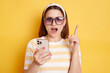 © sementsova321 - Indoor shot of shocked amazed woman wearing striped shirt posing isolated over yellow background, using cell phone, holding smart phone and raised finger up, keeps mouth open.