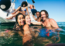 Happy Multiracial Friends Doing Selfie Swimming In The Sea With Sail Boat In Background - Focus On Right Man Face