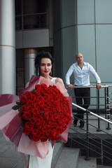 valentine's day. young bride in white wedding dress is standing casual with luxury bouquet of red roses in hands near groom on gray street stairs building background. lifestyle concept, free space