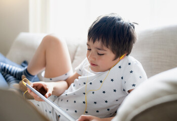 Wall Mural - High key portrait kid wearing earphones listening to music, playing game on tablet,Boy sitting on sofa with light shining from window, Kid playing games on internet,Child doing homework online at home