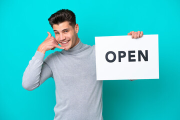 Young handsome caucasian man isolated on blue bakcground holding a placard with text OPEN and doing phone gesture