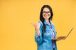 Portrait of happy young beautiful surprised woman with glasses standing with laptop isolated on yellow background. Space for text. Pointing finger.