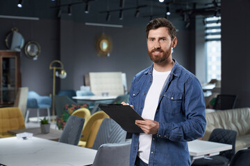 Professional sales assistant in denim shirt standing with dark folder in modern furniture store. Portrait of successful retail manager smiling, while looking at camera in expo center. Concept of work.