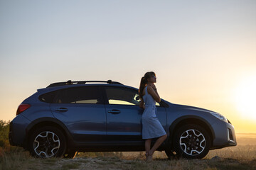 Happy young woman in blue dress standing near her vehicle looking at sunset view of summer nature. Travelling and vacation concept.