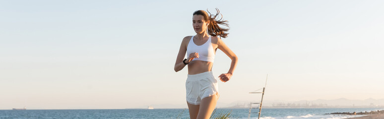 Wall Mural - young woman in shorts and wireless earphone jogging near sea, banner.