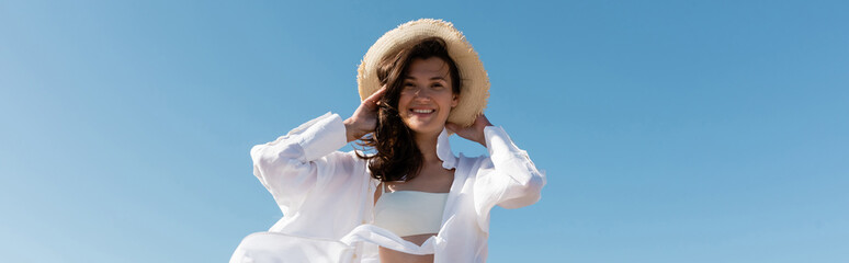 Wall Mural - low angle view of happy young woman in swimsuit and white shirt wearing straw hat against blue sky, banner.